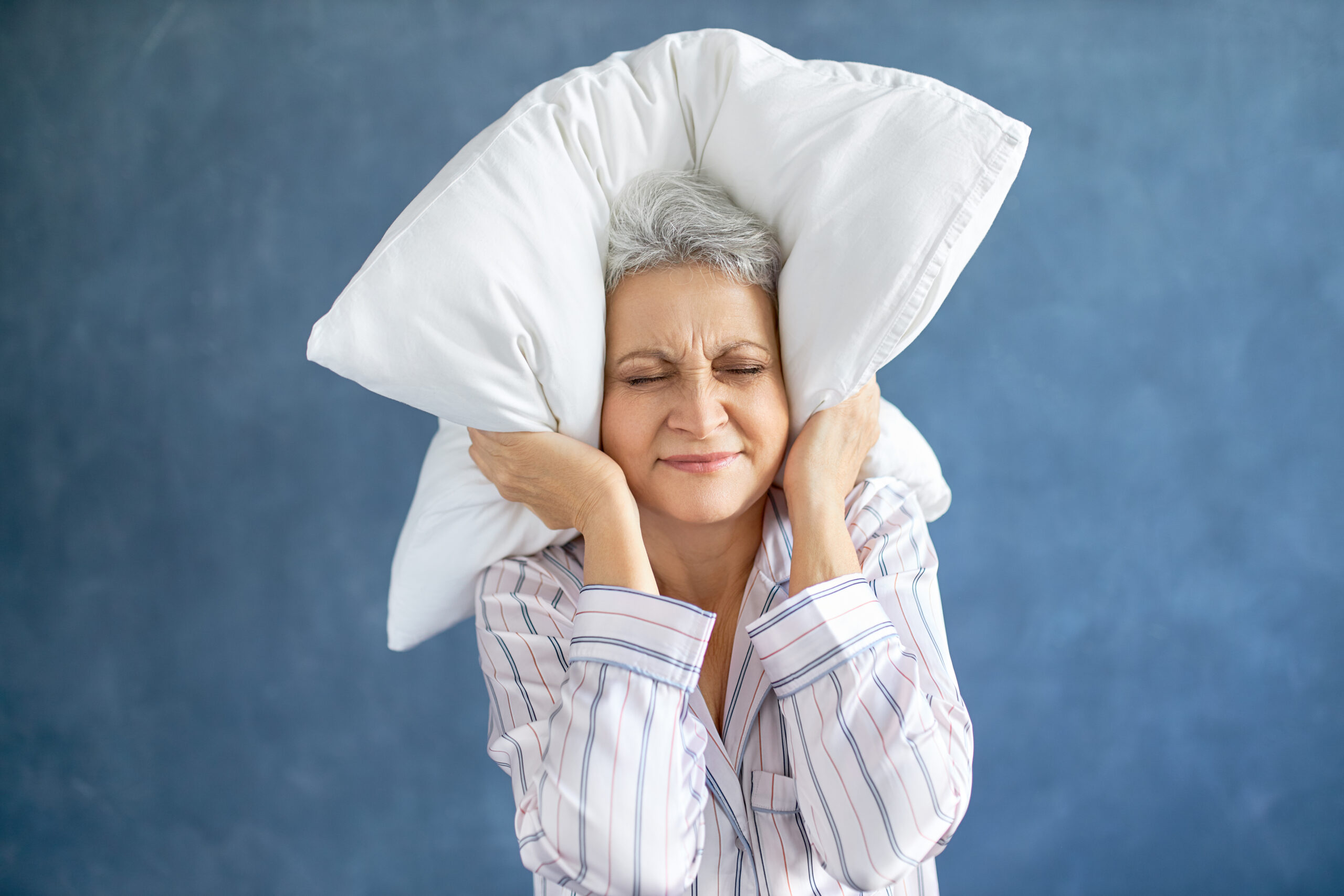 Annoyed dissatisfied mature woman with gray hair grimacing having sleepless night because of loud music, snoring husband, covering ears with pillow, having angry irritated facial expression